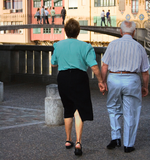 In figure (a), an older man and woman, wearing casual dress, are shown from behind walking in a public plaza setting.
