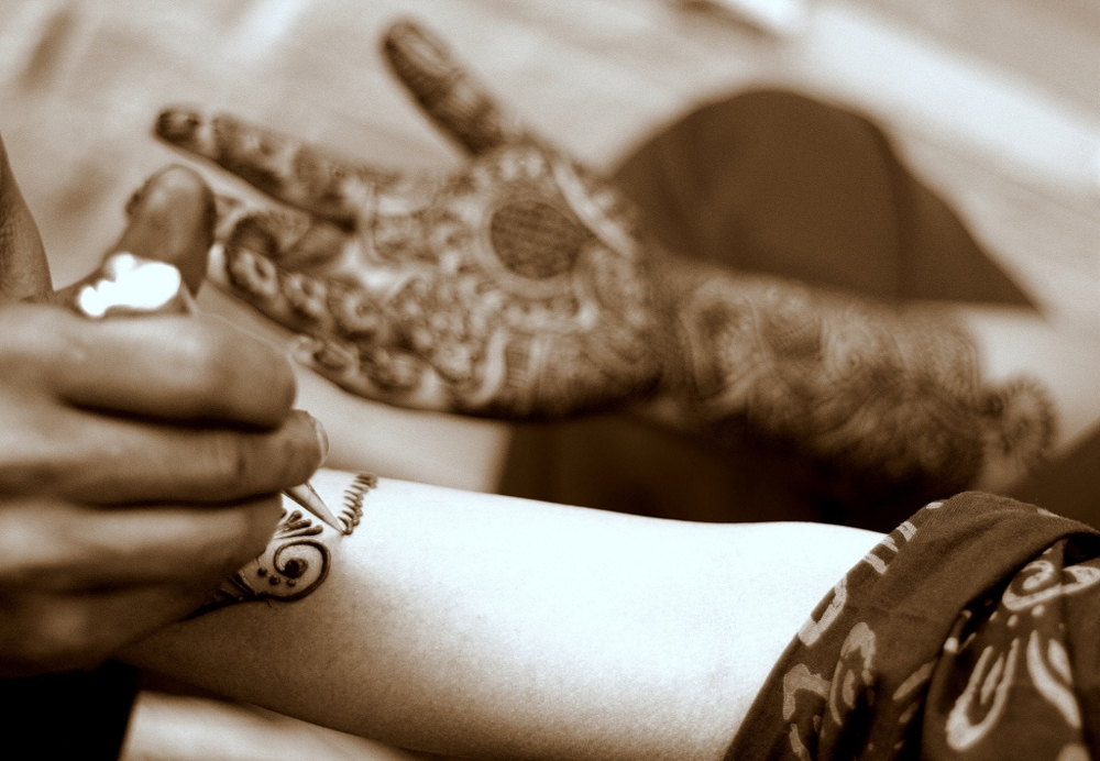 The photo shows a woman’s hands being covered in intricate henna designs.
