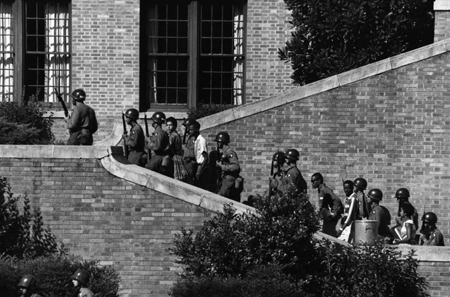 Armed National Guardsmen escorting black students up the outside stairs of a brick high school building.