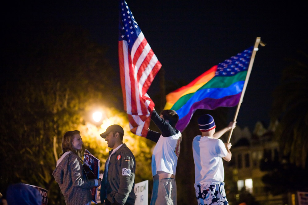 Figure (c) shows people waving a U.S. flag and a rainbow flag.