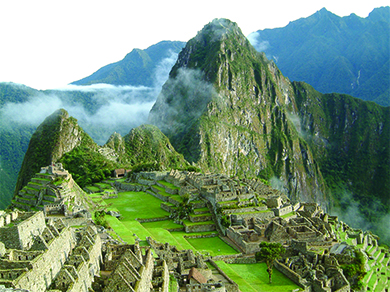 A photograph of Machu Picchu shows the ruins of a complex of buildings with stone walls, stepped terraces green with grass, and a pyramid, with high mountains in the background.