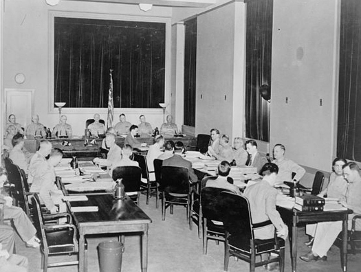 A photo of a group of people in a military commission, seated in chairs around a number of tables arranged in a U shape.