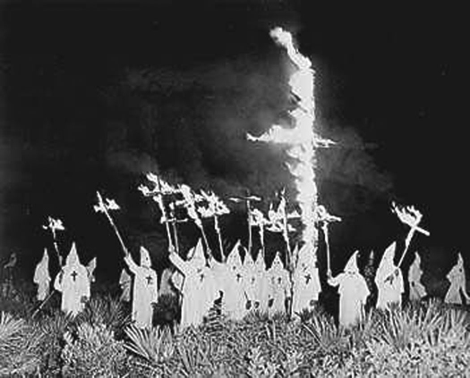 A photo of a group of people wearing robes and pointed hats, surrounding a large cross in the ground that is on fire. Several people hold burning crosses aloft.