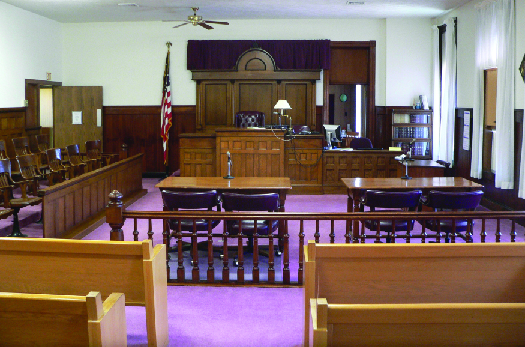 A photo of a typical courtroom, empty of people. In the foreground are benches for attendees, then two tables in the center for the defense and prosecution, and in the background the judge’s stand. To the left of the judge’s stand is a row of chairs for the jury, and to the right of the judge’s stand is the witness stand.
