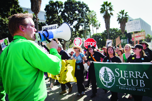 An image of a person speaking through a bullhorn on the left, and a crowd of people marching down a street on the right. Several marchers are holding a large banner that reads “Sierra Club”.