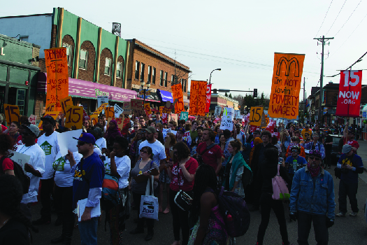 An image of a group of people marching down a street, one of whom holds a sign that reads “I’m not loving’ poverty wages”.