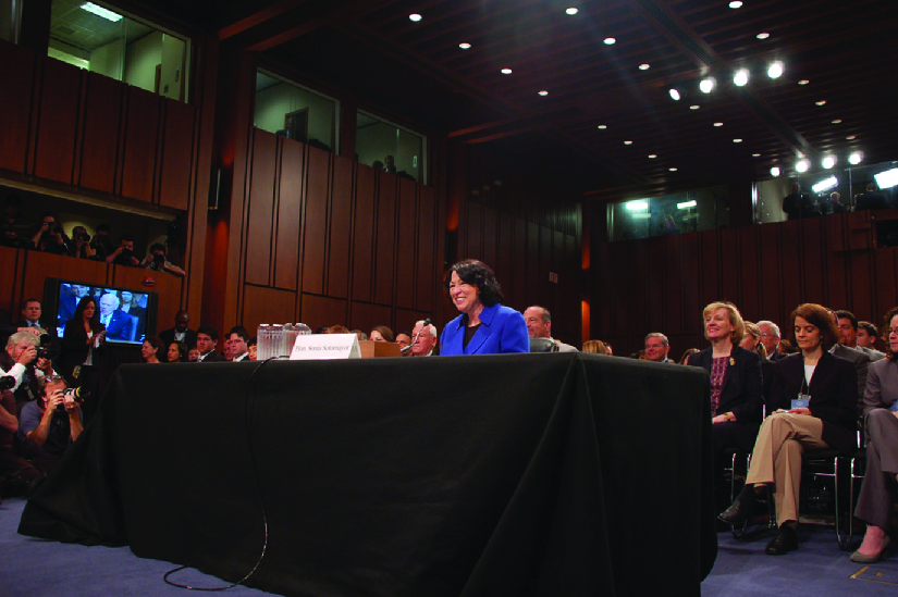 An image of Sonia Sotomayor standing behind a table with a group of people seated behind.