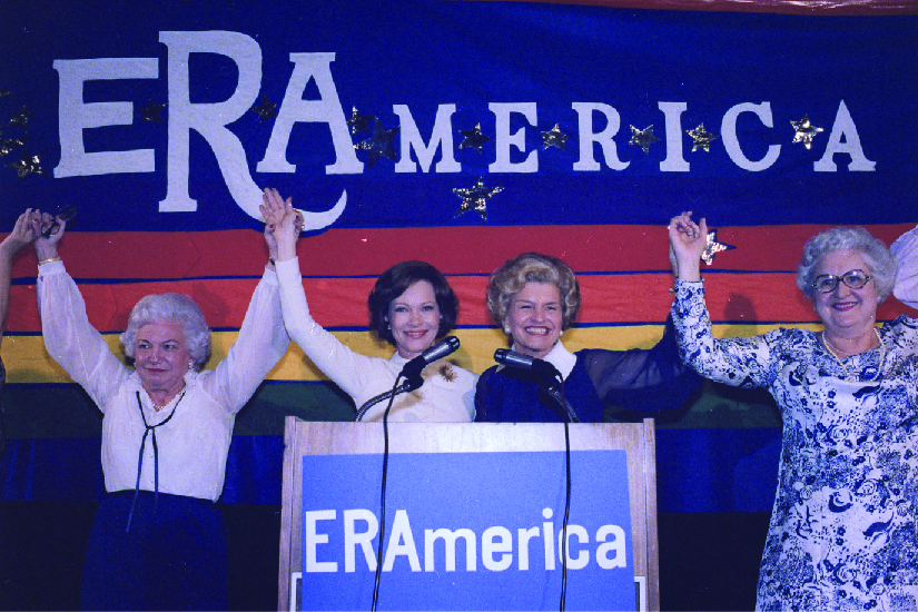 A photo of Rosalynn Carter and Betty Ford speaking at a rally in favor of the Equal Rights Amendment. Signs on stage read “ERAmerica.”