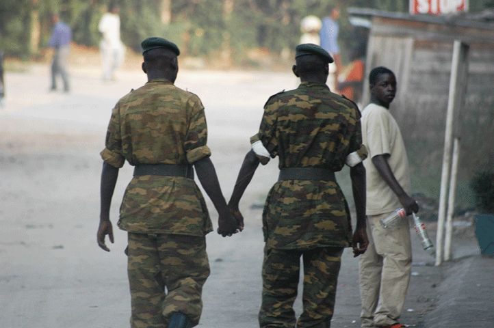 Two male soldiers in uniform are shown from behind walking and holding hands.