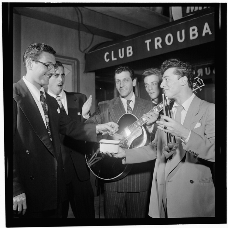 A group of young men wearing suits, including a guitarist, are shown in a black and white photograph in front of the awning of a nightclub.
