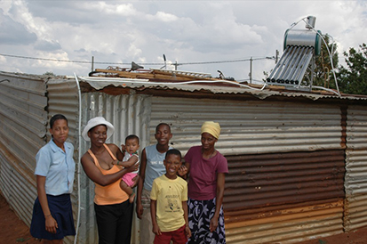 Alt text: A photo of a family of villagers in Africa in front of a solar panel on top of a roof