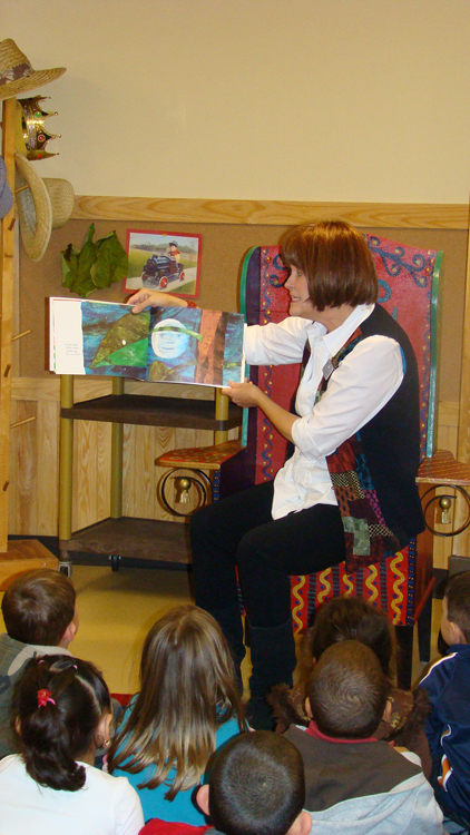 A female teacher is shown sitting in a chair and reading a picture book to a group of children sitting in front of her on the floor.